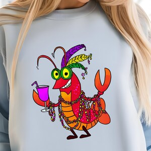 May include: Light blue sweatshirt featuring a cartoon crawfish with a festive Mardi Gras design. The crawfish is red, orange, and yellow, wearing a feathered headdress, beads, and holding a cocktail. The design is a fun, colorful illustration.
