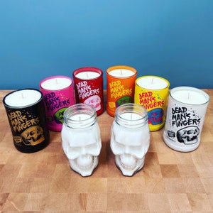 May include: An assortment of candles in various colors, including black, pink, red, orange, yellow, and white. The candles are in glass jars and skull-shaped containers. Each candle has a wick and the text "DEAD MAN'S FINGERS".