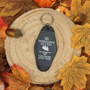 May include: Black key chain with a white motel key tag that says "EST 1693 SANDERSON SISTERS BED & BREAKFAST SALEM, MA CHILDREN STAY FREE". The key tag features a silhouette of three witches riding a broomstick. The key chain is surrounded by autumn leaves and a small orange pumpkin.