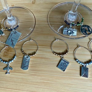 May include: Wine glass charms with travel-themed pendants. Charms include a globe, airplane, postcard, passport, camera, and envelope. Each charm has a beaded ring in silver, gold, or green. The charms are on a wooden surface.