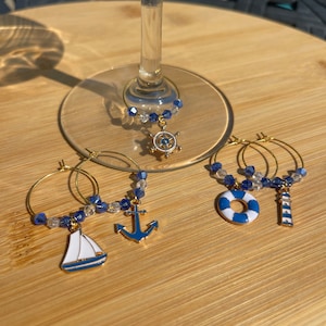 May include: Gold-toned wine glass charms with nautical-themed pendants. Charms include a sailboat, anchor, life preserver, and lighthouse, accented with blue and clear beads. The charms are displayed on a wooden surface.