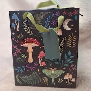Op de afbeelding: Een vierkante zwarte cadeautas met een botanische print van rood-witte paddenstoelen, groene varens, kleine paarse bloemen, een halve maan en een grote groene nachtvlinder. De tas heeft lichtgroene linthengsels en een leeg groen label.