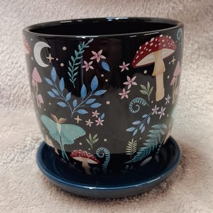Pode incluir: Um vaso de plantas em cerâmica preta com um pires azul-escuro a condizer. O exterior apresenta um padrão colorido de cogumelos com chapéu vermelho, uma traça verde-pálida, luas crescentes brancas, pequenas flores cor-de-rosa e folhagem azul.