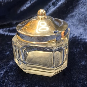 Op de afbeelding: Een heldere, achthoekige glazen pot met een goudkleurig deksel en een kleine, ronde knop. Het deksel heeft een zilveren rand. De pot is waarschijnlijk bedoeld voor het bewaren van suiker, kruiden of andere kleine items.