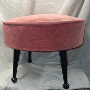 May include: A vintage, round ottoman with a dusty rose velvet upholstered top and sides. The ottoman has four black, tapered legs with rounded feet. The piece is likely from the mid-century modern era.