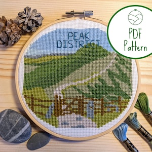 May include: A cross-stitch embroidery hoop featuring a landscape scene of a green hill with a winding path and the text "Peak District". The hoop is on a wooden surface with pine cones and stones.