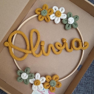 Pode incluir: Um bastidor de bordado circular de madeira com o nome Olivia escrito em fio de malha amarelo mostarda, decorado com seis flores de crochê nas cores amarelo mostarda, branco e verde sálvia. Um pequeno cartão de visita está na caixa.