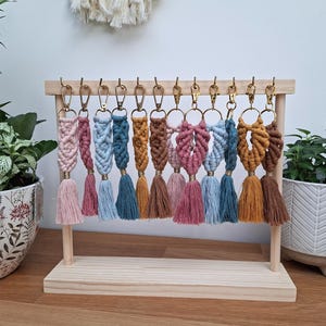 Può includere: Un'esposizione di portachiavi in macramè fatti a mano in vari colori, tra cui rosa, blu e marrone, con elementi metallici color oro. Ogni portachiavi presenta un design intrecciato e un nappa. I portachiavi sono esposti su un supporto di legno.