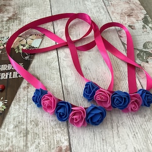 May include: A pink satin ribbon with blue and pink fabric flowers attached. The ribbon is tied in a loop and the flowers are arranged in a cluster.