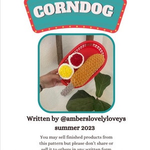 Peut inclure: Un corndog crocheté avec de la moutarde et du ketchup dans un plateau en plastique rouge. L'image comporte le texte "CORNDOG" en haut et "Written by @amberslovelyloveys summer 2023" en bas.