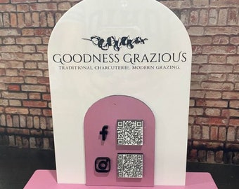May include: A white sign with the text "Goodness Grazious, Traditional Charcuterie, Modern Grazing."  The sign has a pink base and a pink arch with social media icons and QR codes.