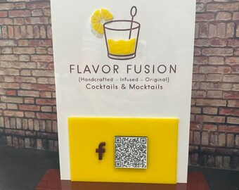 May include: A white sign with the text "FLAVOR FUSION [Handcrafted - Infused - Original] Cocktails & Mocktails" and a yellow graphic of a glass with a lemon wedge and a straw. A yellow square with a Facebook logo and a QR code is attached to the sign.