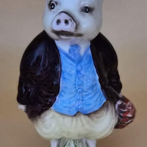 May include: A ceramic pig figurine wearing a blue waistcoat, black jacket, and white trousers. The pig stands on a green base with grass and holds a basket. The figurine is approximately 13 cm tall.