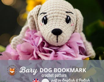 May include: A crocheted dog bookmark, in a light beige color, rests on a bed of pink flowers. The dog has black eyes and a black nose. The text "Bary DOG BOOKMARK crochet pattern pdf in English & Polish" is visible.