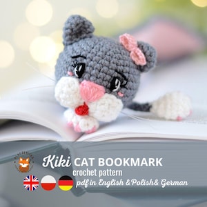 May include: A grey crocheted cat bookmark with pink ears and a pink bow. The cat has white fur on its face and paws. The bookmark is lying on a book with the text "Kiki CAT BOOKMARK crochet pattern pdf in English & Polish & German".