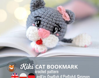 May include: A gray crocheted cat bookmark with pink ears and a pink bow. The cat has white fur on its face and paws. The bookmark is lying on a book with the text "Kiki CAT BOOKMARK crochet pattern pdf in English & Polish & German".