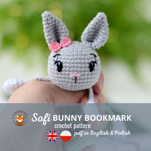 Può includere: Un segnalibro a forma di coniglio all'uncinetto grigio con un fiocco rosa e occhi neri. Il segnalibro è tenuto in una mano. Il testo "Sofi BUNNY BOOKMARK crochet pattern pdf in English & Polish" è sotto il coniglio.