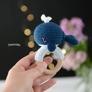 May include: A handmade crochet whale baby rattle in shades of blue and white. The whale has a dark blue body, a white spout, and a light blue ring attached to a natural wood ring. The text "Placzenitka" is on the left side.