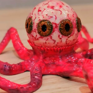 Peut inclure: Une sculpture de poulpe faite à la main, avec une tête tachetée de blanc et de rouge, ornée de plusieurs yeux bruns et verts. Les tentacules et le corps sont d'un rouge vif, créant une esthétique saisissante et troublante. Sculpture unique.