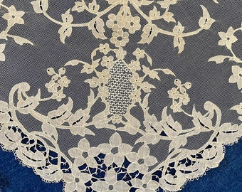 May include: A delicate, off-white lace collar with a floral design. The lace is intricate and features a central motif of a small, oval shape surrounded by leaves and flowers.