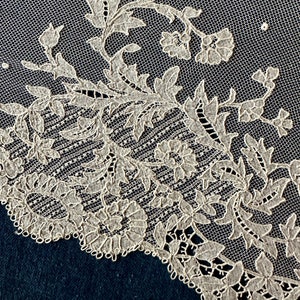 May include: Close-up of a delicate white lace fabric with a floral pattern. The lace features intricate details and a scalloped edge.