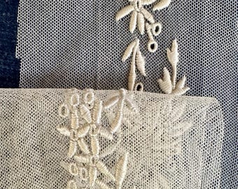 May include: Close-up of ivory-colored lace with floral embroidery. The delicate design features embroidered flowers, leaves, and small berries. The lace has a fine, open weave, and the embroidery adds texture and detail to the fabric.