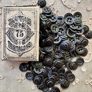 May include: A collection of dark green buttons scattered around a vintage cardboard box. The box is labeled "BOUTONS POUR PANTALONS" with the number 75. The buttons are round with four holes, and the box is tied with twine. The background is a beige lace fabric.