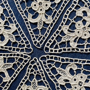 Pode incluir: Seis guardanapos de renda branca em forma triangular com intrincados desenhos florais. Os guardanapos estão dispostos em um padrão circular sobre um fundo azul escuro.