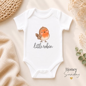 May include: A white short-sleeved baby bodysuit featuring a central graphic of a small, round, brown and orange robin bird with a smiling expression. Below the bird, the text little robin is printed in a black, cursive, lowercase font.
