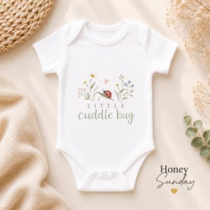 Puede incluir: Un body blanco de manga corta para bebé con un gráfico central de una mariquita roja sobre una hoja verde, rodeada de pequeñas flores silvestres y un pequeño corazón. El texto "LITTLE cuddle bug" está impreso en color verde oscuro.
