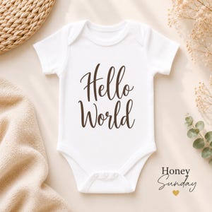 Op de afbeelding: Een witte babybody met korte mouwen en de tekst Hello World in een donkerbruin cursief lettertype. Het kledingstuk heeft een ronde halslijn en drie drukknopen aan de onderkant. Het ontwerp is gecentreerd op de voorzijde van de witte stof.