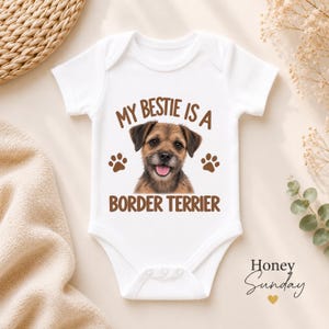 May include: A white short-sleeved baby bodysuit featuring a graphic of a brown Border Terrier dog with its tongue out. The text reads "MY BESTIE IS A BORDER TERRIER" in brown letters, flanked by two brown paw prints on either side of the dog.