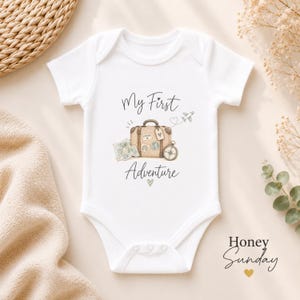 May include: A white short-sleeved baby bodysuit featuring a graphic illustration of a brown suitcase, a map, a compass, and a small aeroplane. The text reads My First Adventure in a dark script font, with a small heart icon below the word Adventure.