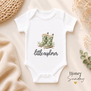 May include: A white short-sleeved baby bodysuit featuring a graphic of two sage green rain boots with white star patterns and mud splatters. Below the boots, the text reads little explorer in a black script font. The garment has a round neckline.