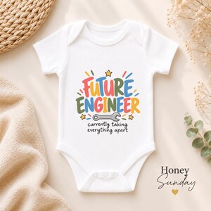 Peut inclure: Un body blanc à manches courtes pour bébé avec un motif coloré portant l'inscription Future Engineer en lettres rouges, vertes et bleues. Sous le texte se trouve une illustration de clé grise et le texte noir currently taking everything apart.
