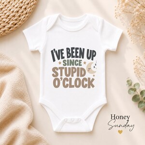 May include: A white short-sleeved baby bodysuit featuring the text I'VE BEEN UP SINCE STUPID O'CLOCK in dark gray, green, and brown lettering. The design includes a small crescent moon with closed eyes and three Zs, plus two small stars.