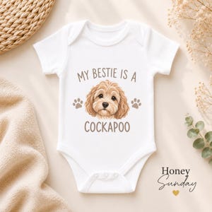 Pode incluir: Um body branco de manga curta para bebé com um gráfico castanho da cara de um cão Cockapoo no centro. O texto acima do cão diz MY BESTIE IS A e o texto abaixo diz COCKAPOO, ladeado por dois pequenos ícones de pegadas castanhas.