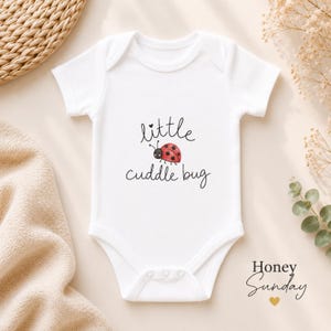 Op de afbeelding: Een wit babyrompertje met korte mouwen en een aquarelillustratie van een rood lieveheersbeestje met zwarte stippen. De tekst "little cuddle bug" is in zwarte schrijfletters boven en onder het lieveheersbeestje gedrukt.