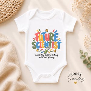 May include: A white short-sleeved baby bodysuit featuring a colourful graphic that reads FUTURE SCIENTIST in bold, multi-coloured letters, accompanied by a green test tube illustration and the text currently experimenting with everything.