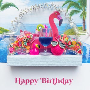 May include: A birthday card with a tropical theme, featuring a pink flamingo, a glass of blue liquid, and a pink pom-pom wearing sunglasses. The card includes star-shaped containers filled with fruit decorations and the words "Happy Birthday".