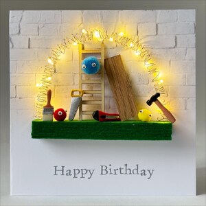 May include: A handmade birthday card with a construction theme. It features miniature tools, a ladder, and a wooden plank set against a white brick wall. The words "Happy Birthday" are printed at the bottom, and fairy lights are strung above the scene.