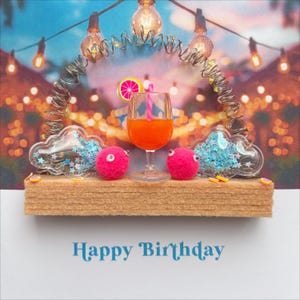 May include: A whimsical birthday card featuring a miniature cocktail in a glass, flanked by two pink pom-poms with googly eyes and cloud-shaped decorations filled with blue glitter. The card has the words "Happy Birthday" in blue.