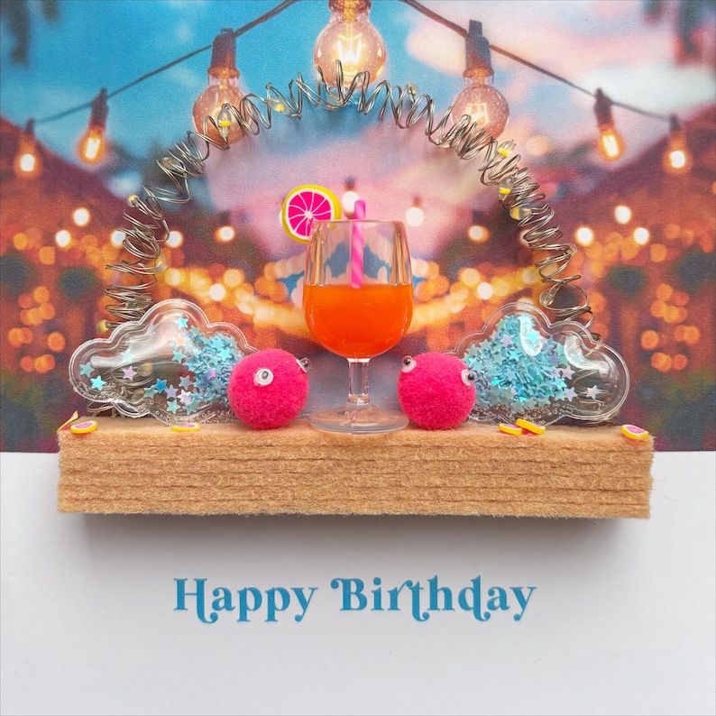 May include: A whimsical birthday card featuring a miniature cocktail in a glass, flanked by two pink pom-poms with googly eyes and cloud-shaped decorations filled with blue glitter. The card has the words "Happy Birthday" in blue.