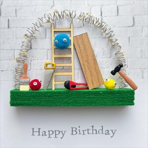May include: A handmade birthday card with a construction theme. It features a wooden ladder, a saw, a hammer, a paintbrush, and a piece of wood. The card has a green base and a white brick background. The text "Happy Birthday" is printed at the bottom.
