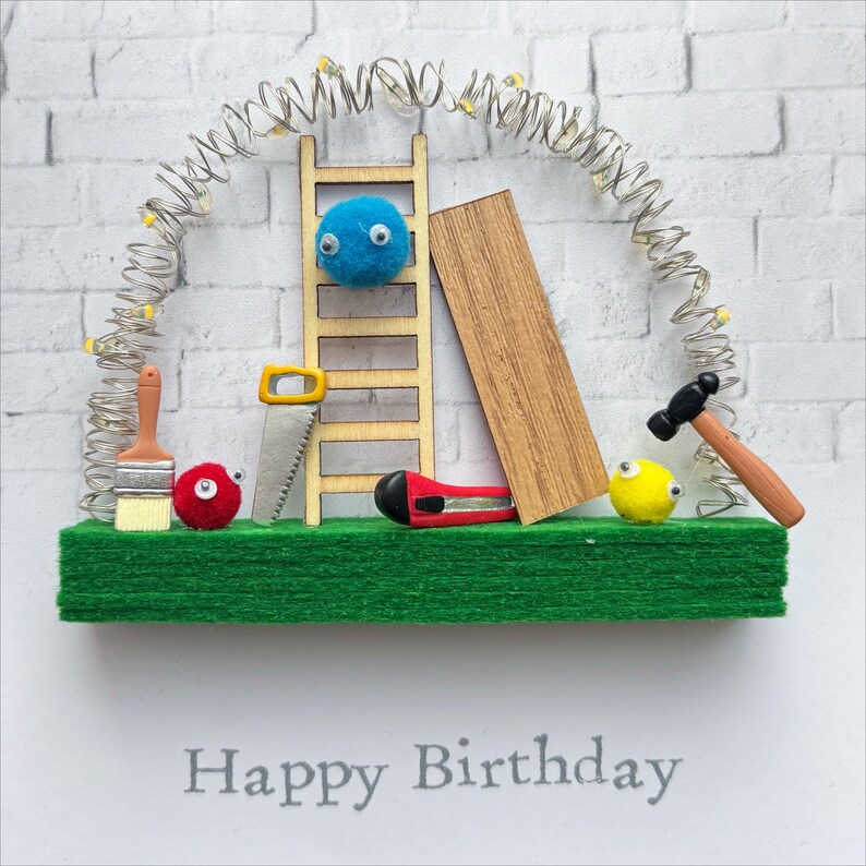 May include: A handmade birthday card with a construction theme. It features a wooden ladder, a saw, a hammer, a paintbrush, and a piece of wood. The card has a green base and a white brick background. The text "Happy Birthday" is printed at the bottom.