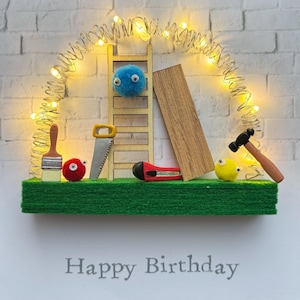 May include: A whimsical birthday card featuring a miniature construction scene. A wooden ladder, saw, and other tools are arranged on a green base, with a string of lights overhead. The words "Happy Birthday" are printed at the bottom.