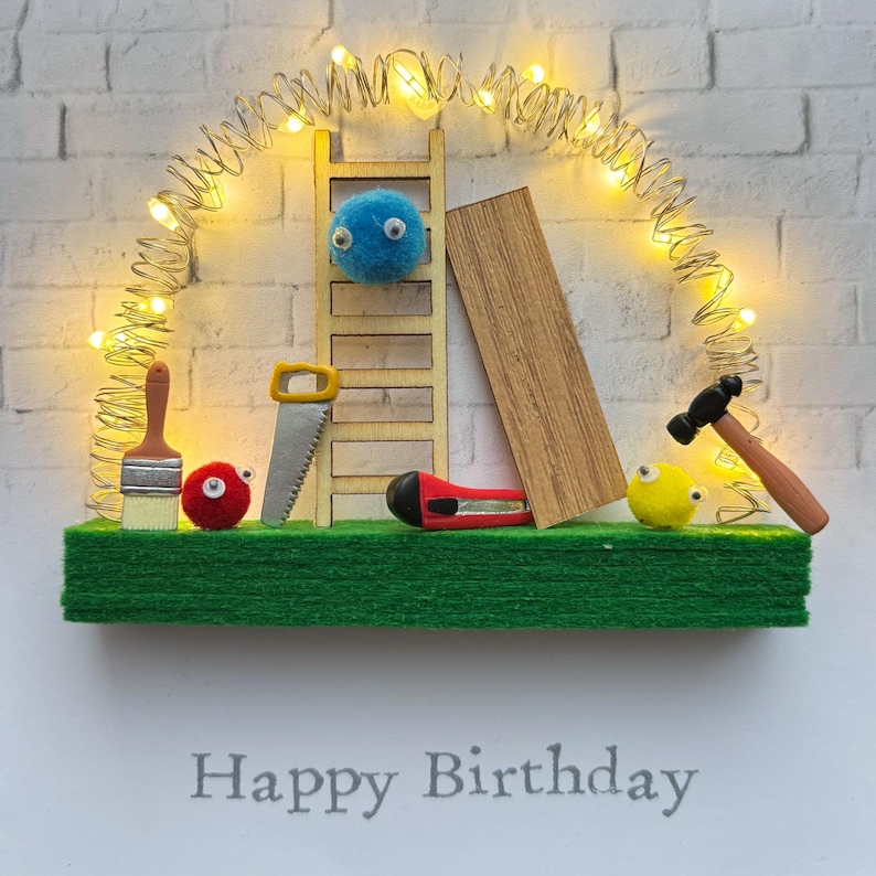 May include: A whimsical birthday card featuring a miniature construction scene. A wooden ladder, saw, and other tools are arranged on a green base, with a string of lights overhead. The words "Happy Birthday" are printed at the bottom.