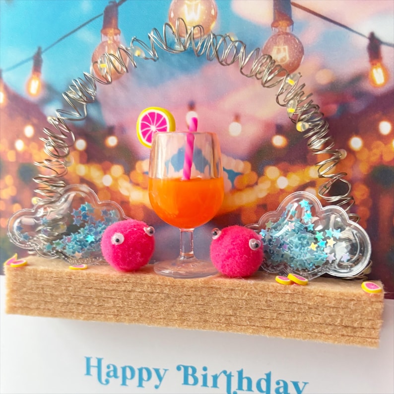 May include: A birthday card featuring a cocktail in a glass, two pink pom-pom creatures, and cloud-shaped containers with blue stars. The card has the words "Happy Birthday" at the bottom, with a background of string lights.