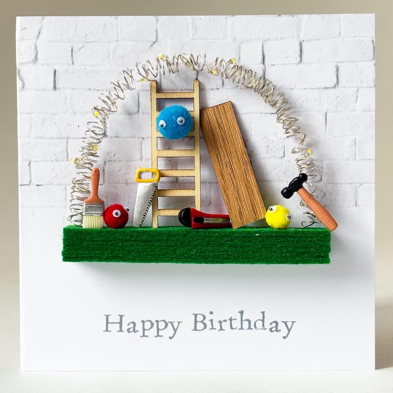 May include: A handmade birthday card with a construction theme. It features a wooden ladder, a piece of wood, and various tools like a hammer and saw, all set against a white brick wall. The words "Happy Birthday" are printed at the bottom.
