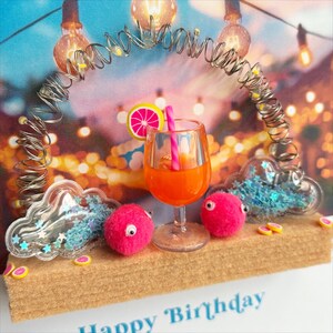 May include: A decorative birthday card featuring a cocktail in a glass with a pink straw and a citrus slice. The card includes pink pom-poms with googly eyes, cloud-shaped containers with blue glitter, and a metal arch with lights. The words "Happy Birthday" are visible.